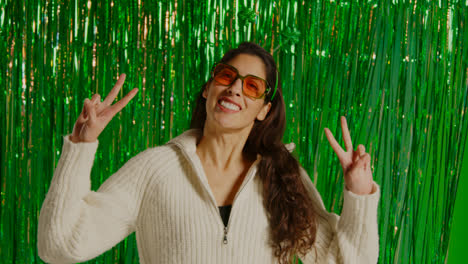 Woman-Celebrating-St-Patrick's-Day-Standing-In-Front-Of-Green-Tinsel-Curtain-Wearing-Prop-Shamrock-Deely-Boppers-And-Glasses